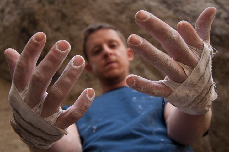 Lifestyle_Tommy_hands_(c)BeccaCaldwell_DawnWall Tommy Caldwell wears a blue shirt and looks down at his hands that are the central focus of the shot. His hands are covered in chalk and tape.