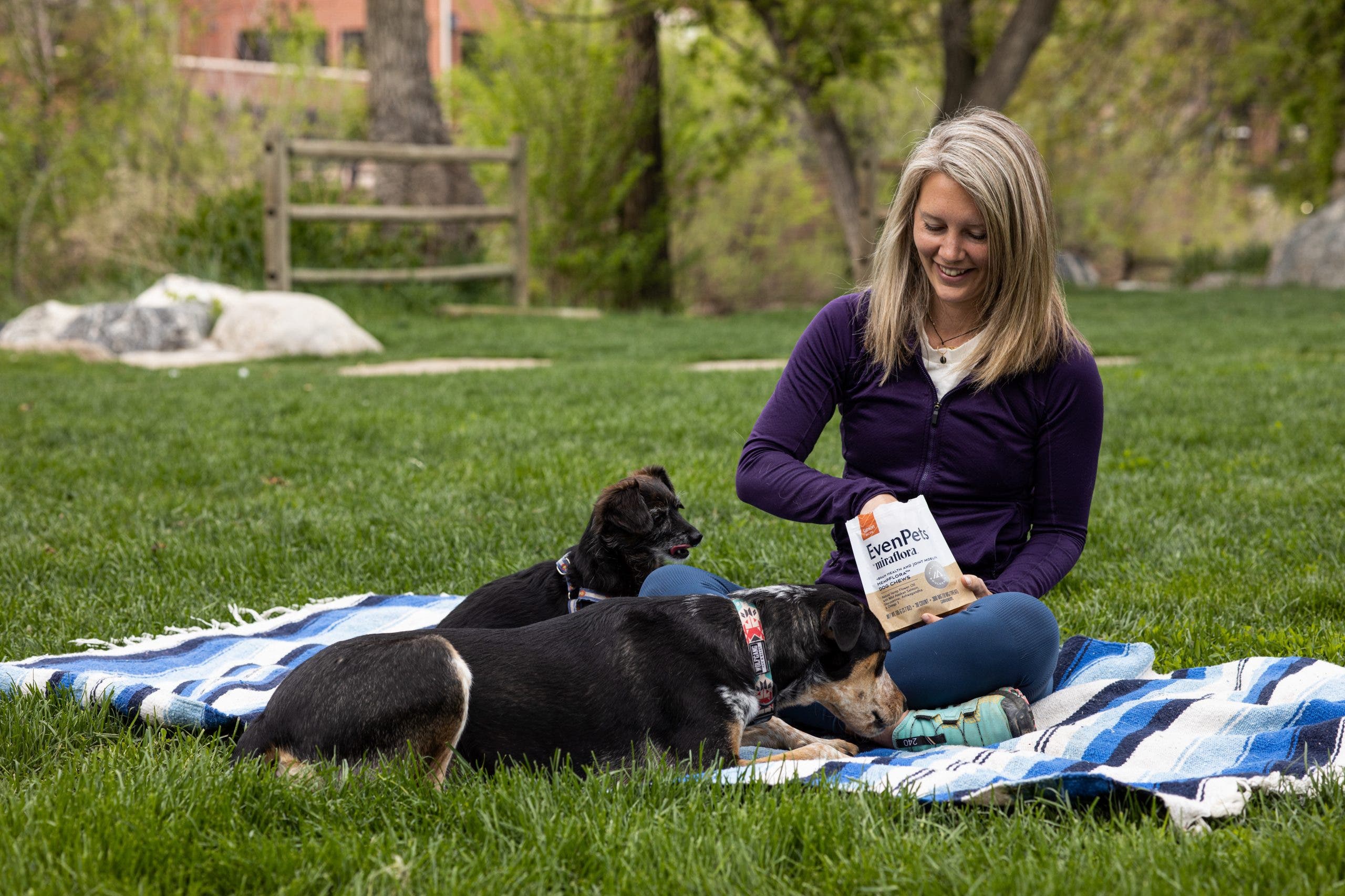 Chelsea Rude sits on a patterned blue blanket in the grass in Golden, CO with her two dogs, feeding them CBD treats.