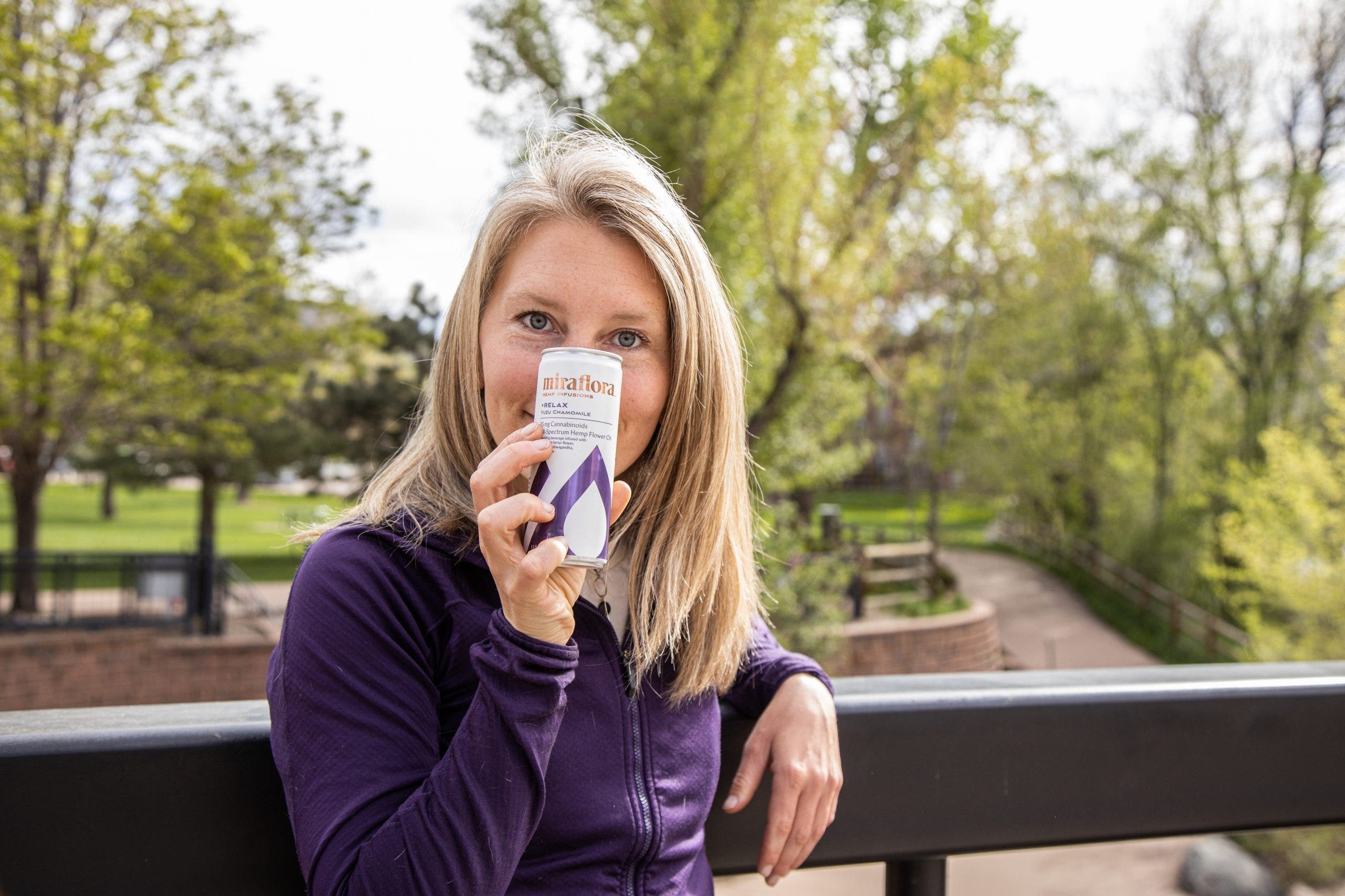Chelsea Rude poses with a can of sparkling CBD beverage next to a park. She is smiling and her purple jacket matches the purple can.