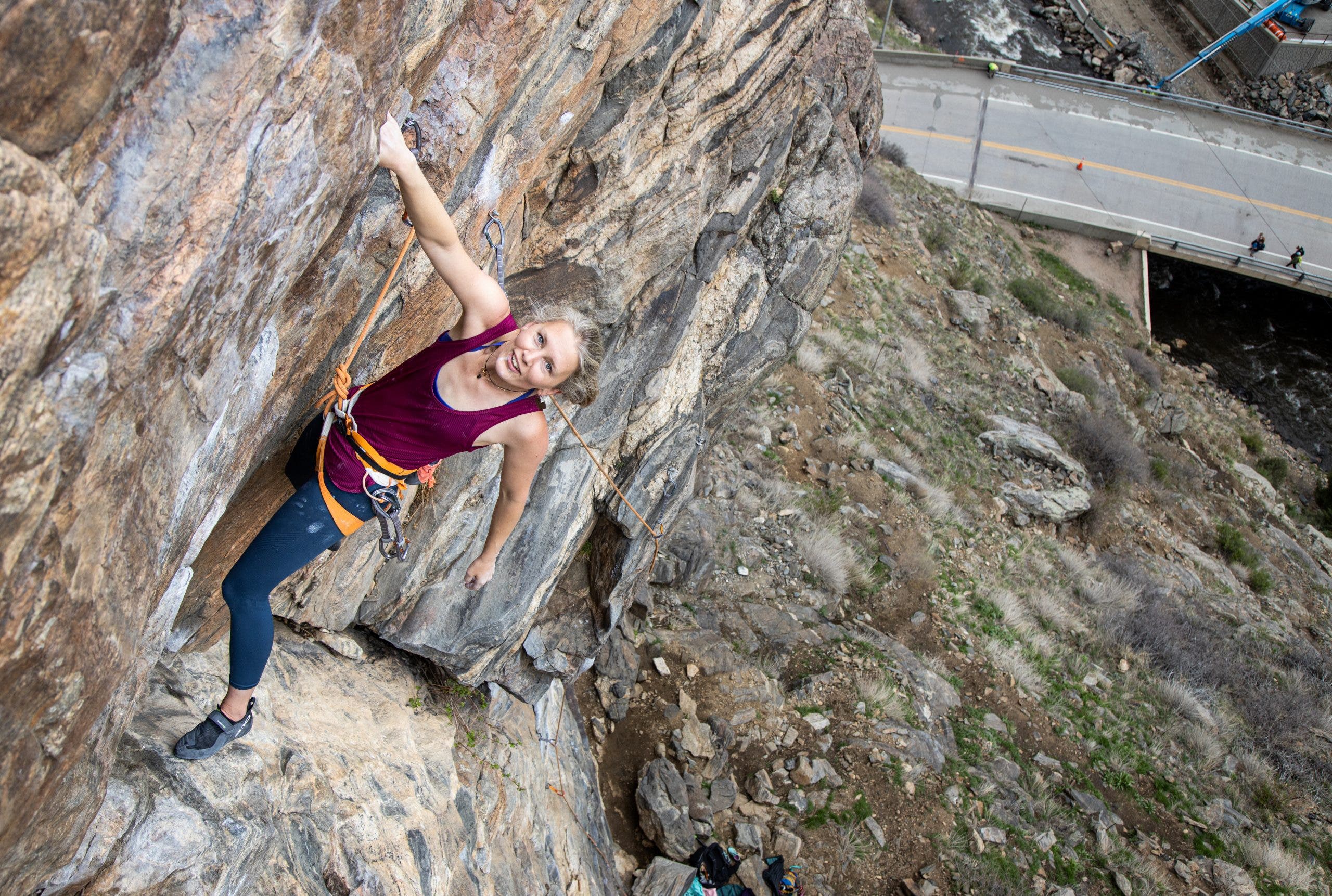 Chelsea Rude climbs on a bright orange rope up a route in Clear Creek Canyon in a pink tank top and blue leggings. She is smiling at the camera, this was one of her first climbing days post injury recovery.