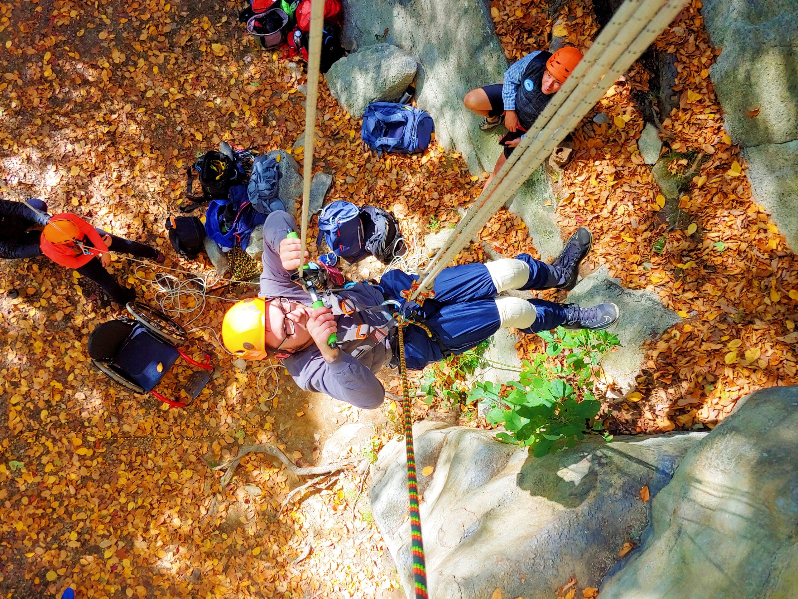 A man ascending a set of fixed ropes without the use of his legs. The photo is top down. The man is wearing a yellow helmet. The ground is coated with autumn-colored leaves, yellow, orange, and amber.