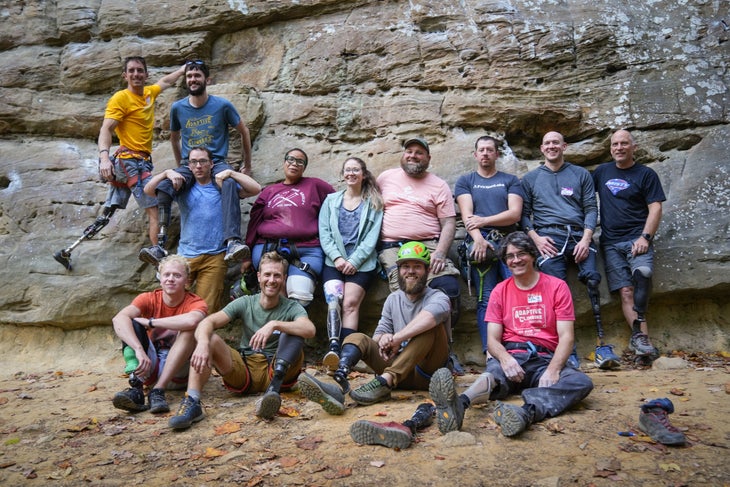 Athletes at the climbing clinic for lower limb amputees gather at the base of the cliff for a group photo. Smiling and tired after an afternoon of climbing they all wear prosthetic legs, fitted with either climbing shoes or hiking boots. From left to right: Ronnie Dickson, Colin Torpey, Kyle Long, Kareemah Batts, Kara Parker, Jeffrey Stiner, Jeremy Ritchie, Ryan Hurd, Jim Ewing, Jono Lewis, Tanner Jones, and John Freeland,
