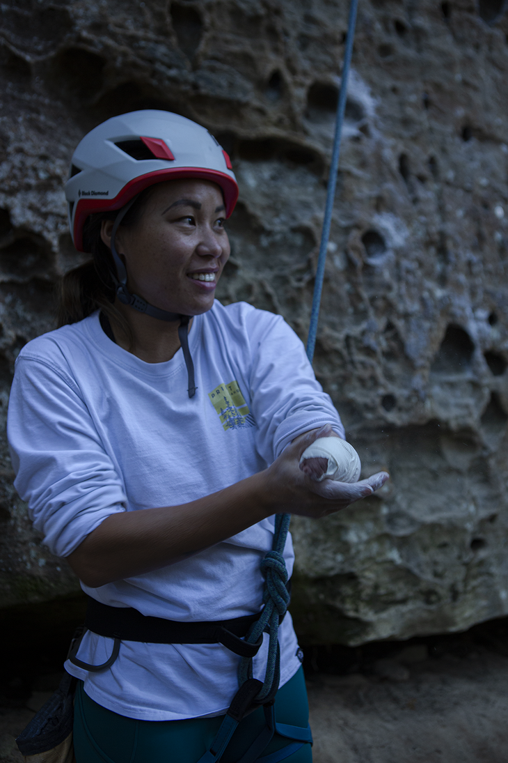 Leah Kaplan is tied into the end of a blue rope. She stands in front of a wall of chalky pockets wearing a white helmet with white tape around her nub, and a smile.