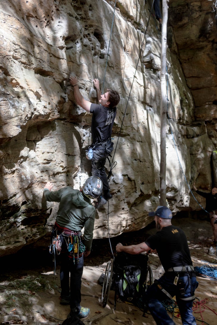 Ascending above his wheelchair with a series of campus moves, Tanner Cislaw holds confidently to the sandstone face. A volunteer moves his chair out of the way as Tanner climbs up the wall.