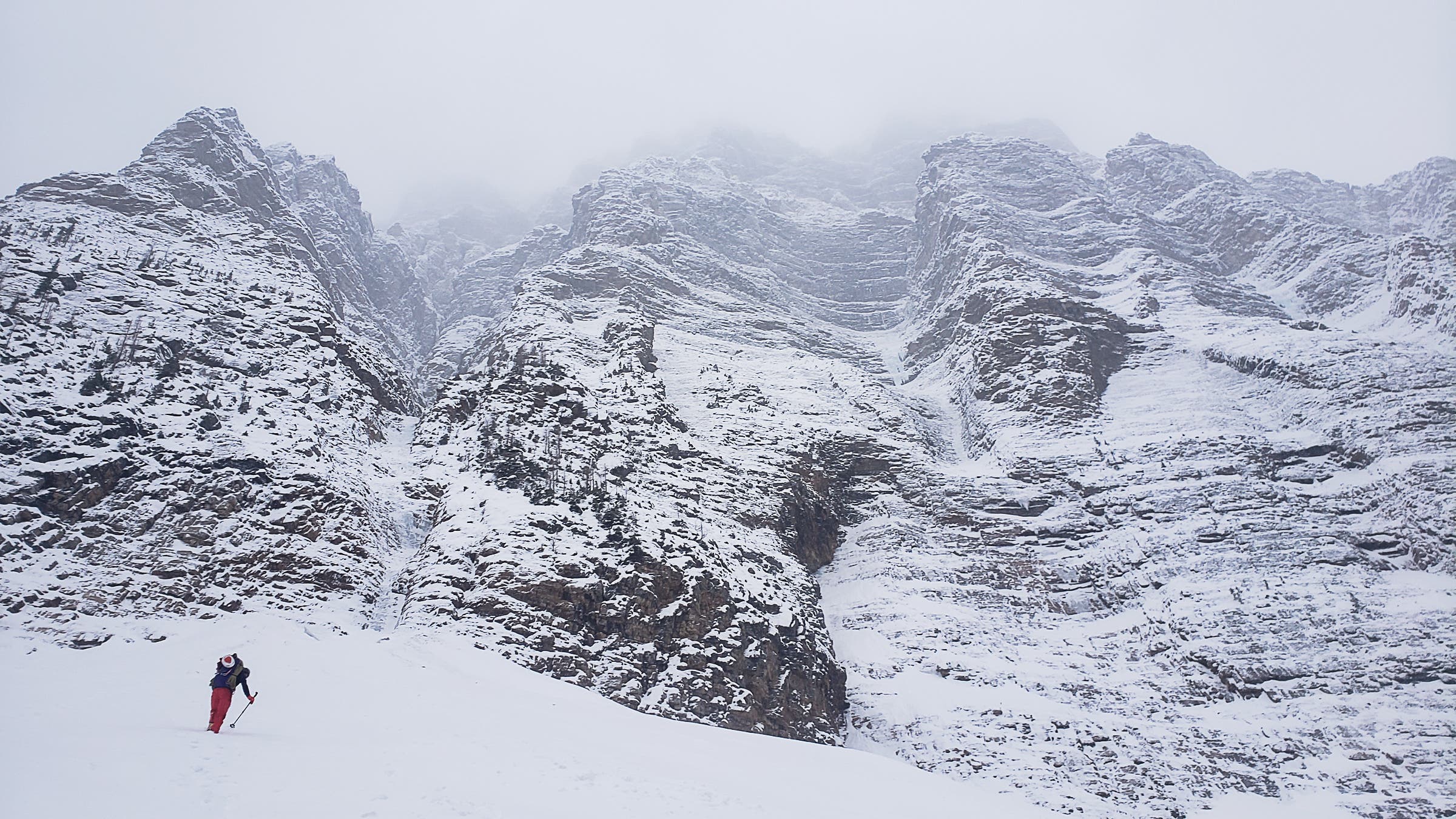 Man walks up snow slope beneath tall, rocky mountain.