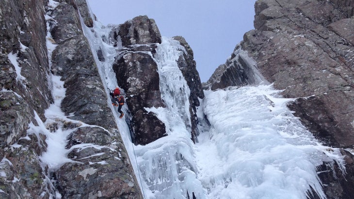 Ice climber ascends vertical ice pitch surrounded up steep rock.