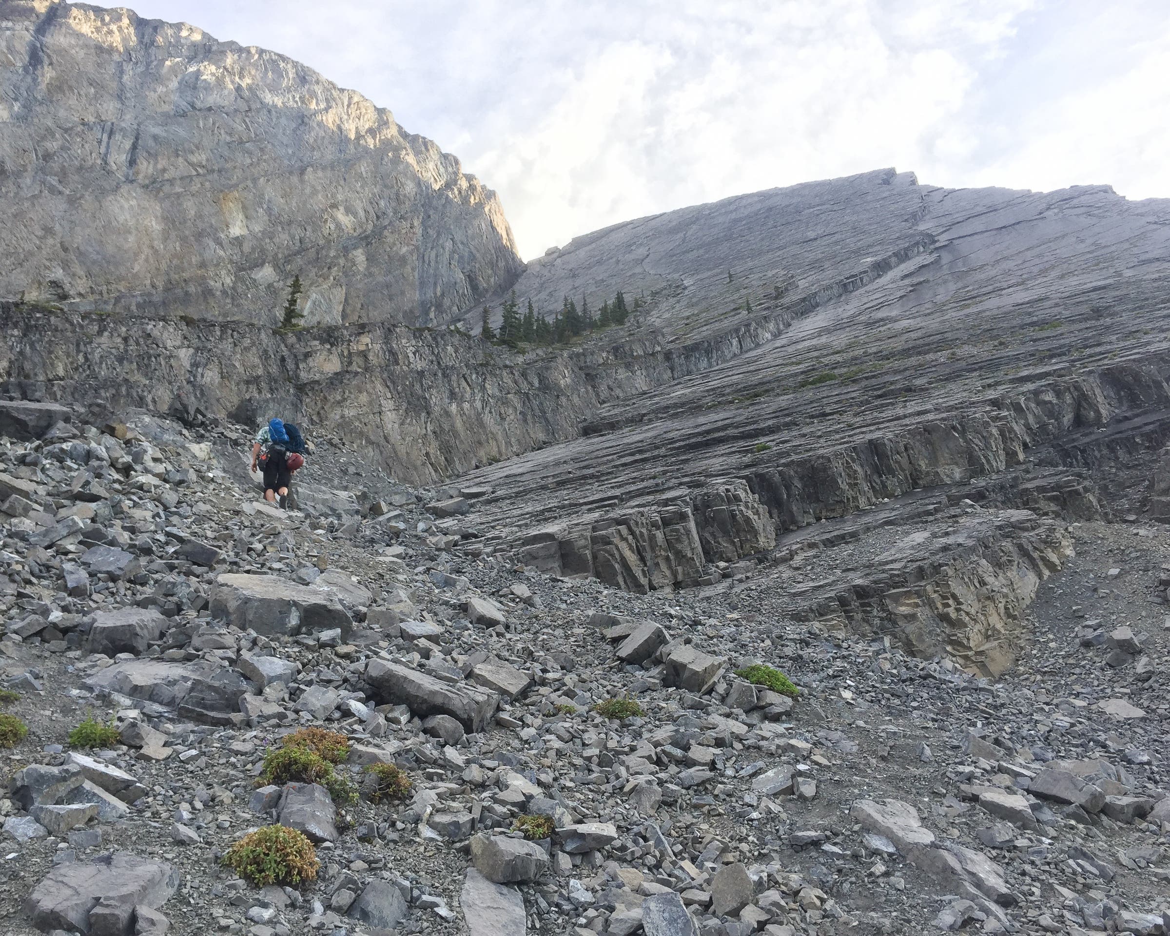 Hiker walks below slabby mountain face on a cloudy day.