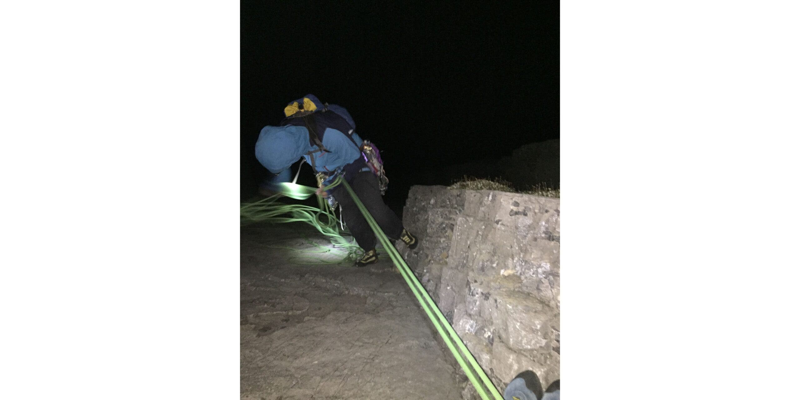 Man in blue jacket rappels down rocky corner at night, illuminated by headlamp.