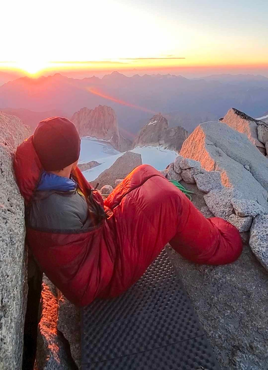 A climber sits upright in his sleeping bag on the summit of a tall mountain at sunrise.