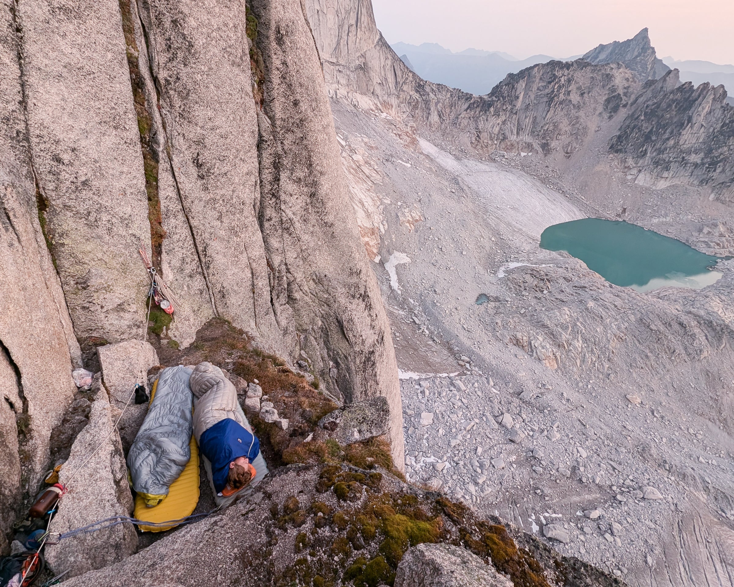 A climber sleeps on an exposed ledge partway up a steep mountain.