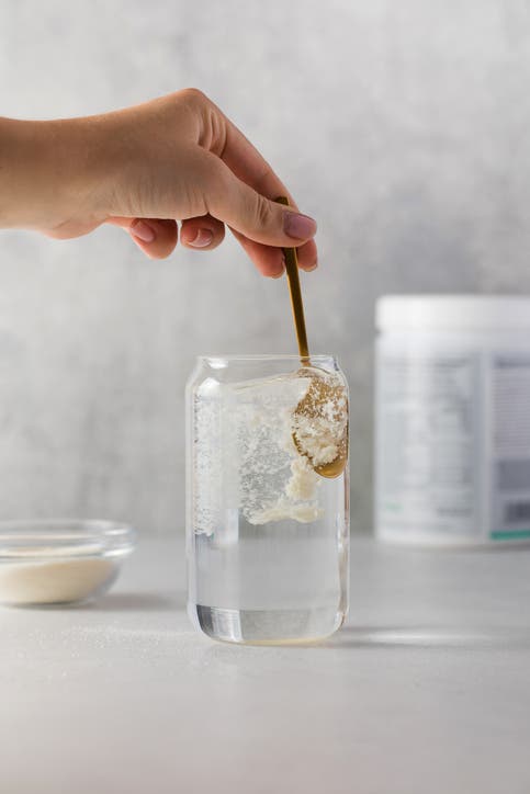 Hydrolyzed collagen powder is added with a spoon to a transparent glass of water on a grey background.