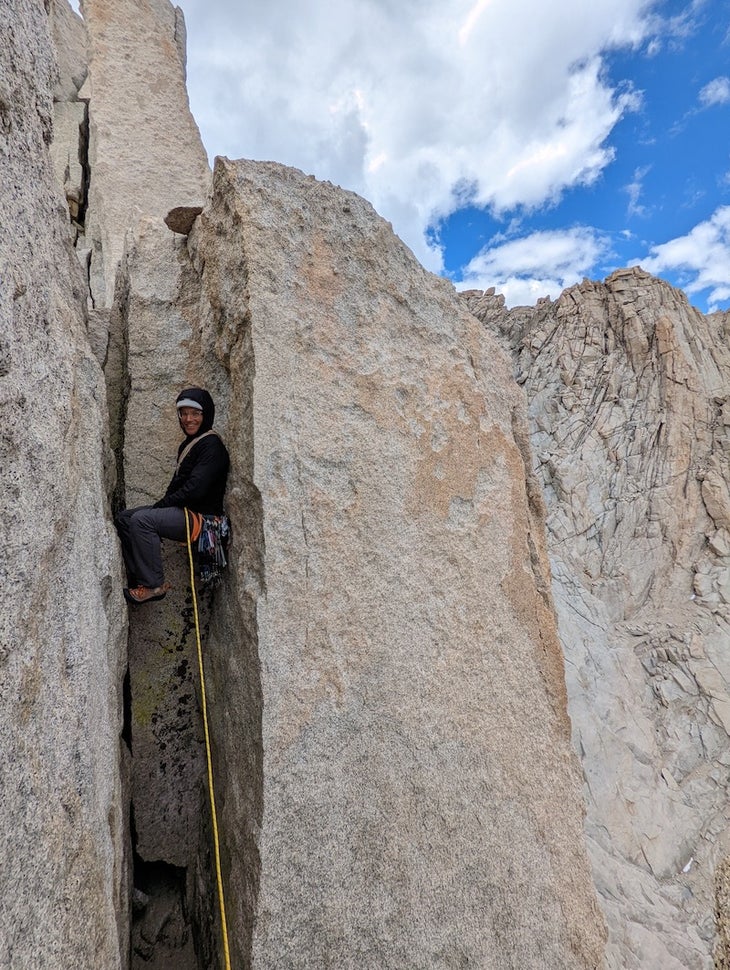 Adam Shmidt chimneying up a granite cliff, quite runout, smiling over at the camera.