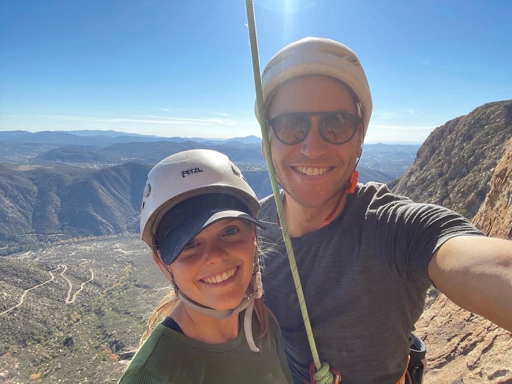 A selfie of a smiling couple high on a California rock climb–the desert and the sky behind them.