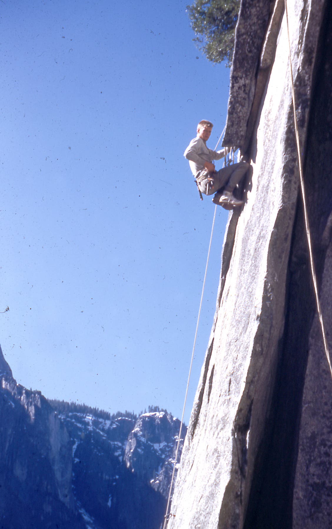 Man climbs a vertical pane of granite on a sunny day in Yosemite, snow dusting trees in background.
