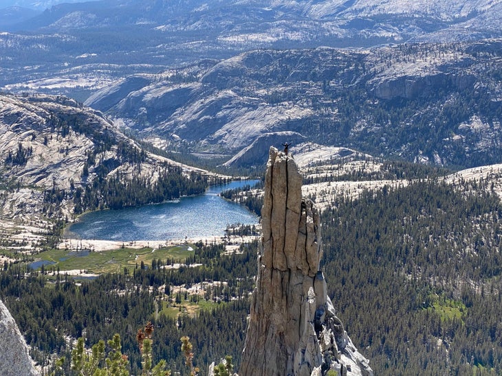 A figure standing on a thin granite tower in the Yosemite high country, arms spread high, with a shiny blue lake and rocky hills in the distance.