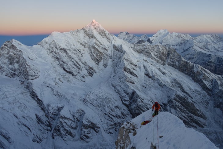 Climber stands on exposed snow ridge at sunset on Himalayan peak.