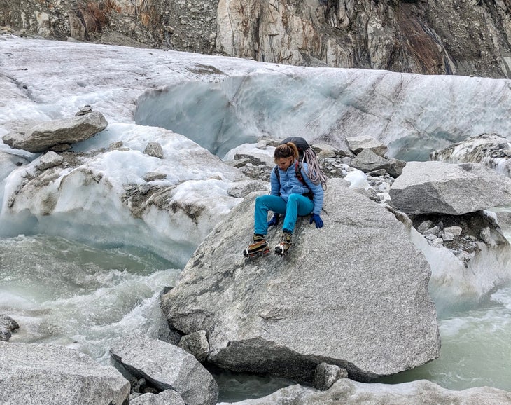 A woman climber wearing crampons crossing a river via a big boulder.