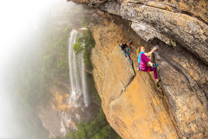 Man and woman climb steep rocky cliff beside waterfall in Australia.