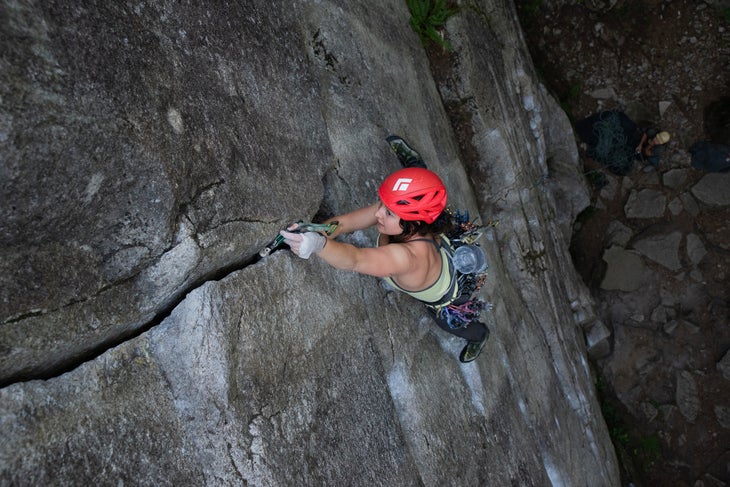 Female climber wearing red helmet placing cam in crack on rock wall.