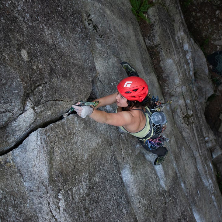 Female climber wearing red helmet placing cam in crack on rock wall.