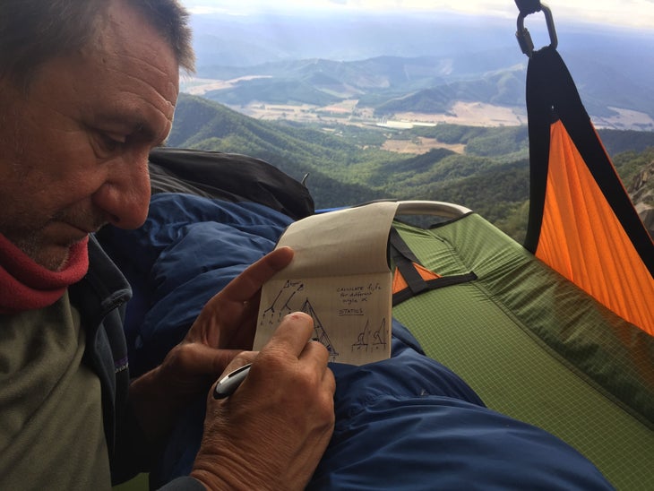 Middendorf lying in a portaledge on a big wall while designing a portaledge on a notepad—calculating angles and writing notes.
