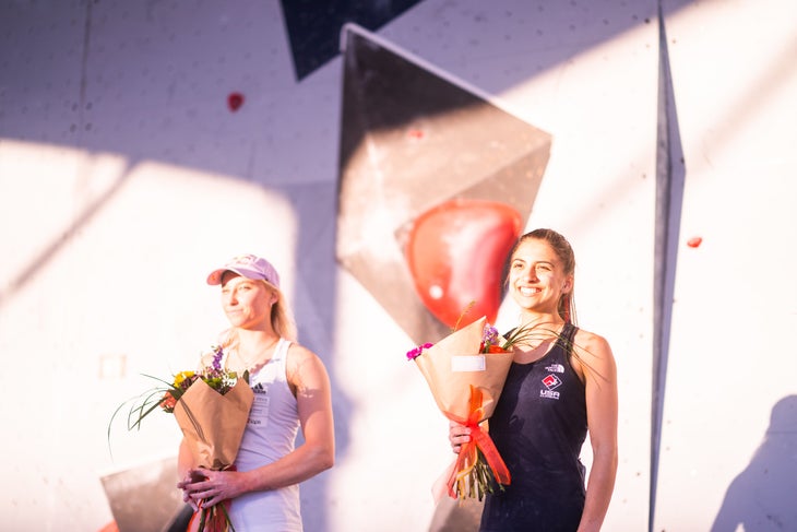 Two female competition climbing rivals—Janja Garnbret and Natalia Grossman–holding flowers under the spotlights in front of a bouldering wall.