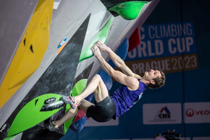 Adam Ondra grimacing as he sets up for a powerful move on green pinches during a bouldering final.