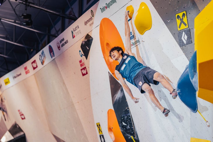 A member of Team Japan hanging one handed from the finishing hold of a World Cup Boulder. He's smiling.