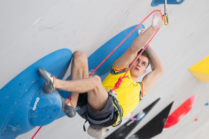 A male competition climber demonstrating a rose move (one of our comp climbing terms) while clipping.