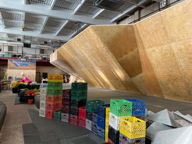 An image of bare climbing walls with boxes full of climbing holds in the foreground. In the background, a sign saying: in GP81 we trust.