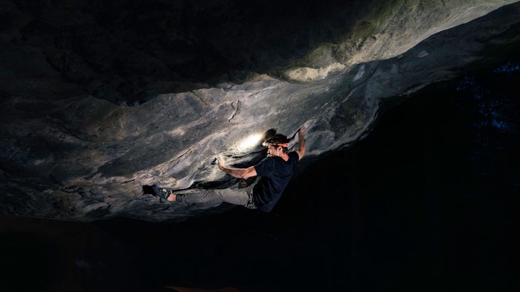 A night bouldering photograph of Sam Weir (wearing a headlamp) doing a very hard sequence on a shadowy, overhanging boulder.