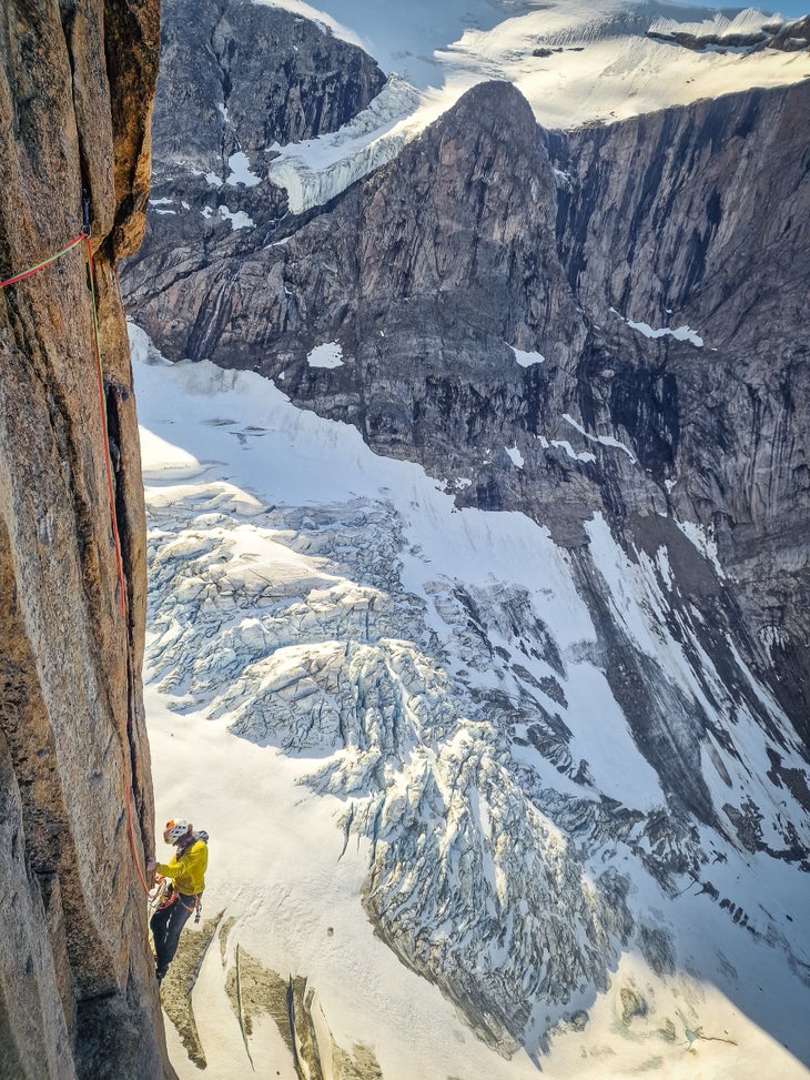 Man rock climbers a vertical granite crack on a mountain with glaciers behind him.