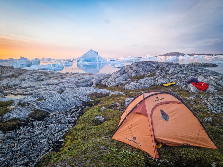 A pink tent is set up at sunset in the arctic, on the edge of an iceberg-filled fjord.