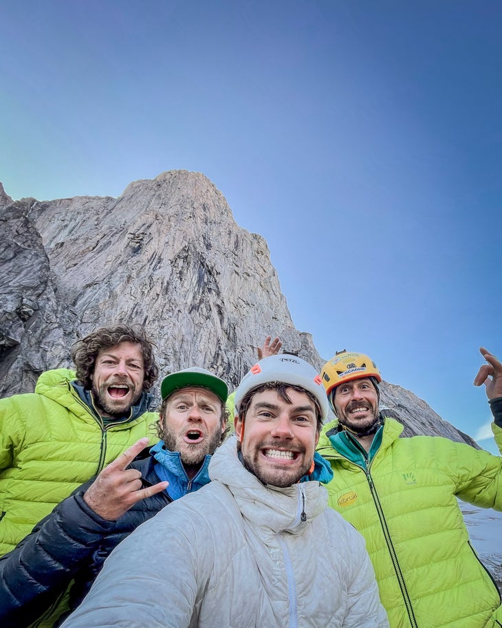 Four bearded men smile beneath a big mountain in Greenland on a sunny day.