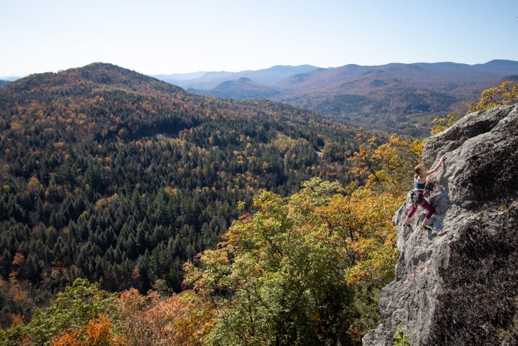 A woman climbing near the top of a cliff with excellent autumnal views of the White Mountains behind her.