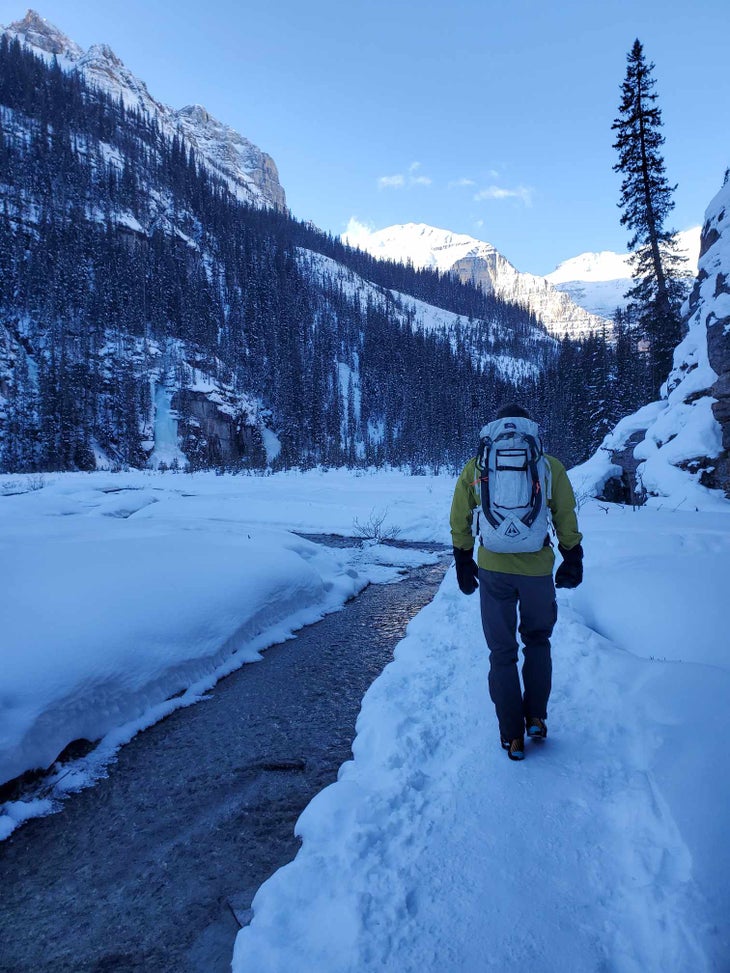 Man hikes in wintery mountain valley with Black Diamond Hydra ice tools attached to his backpack.