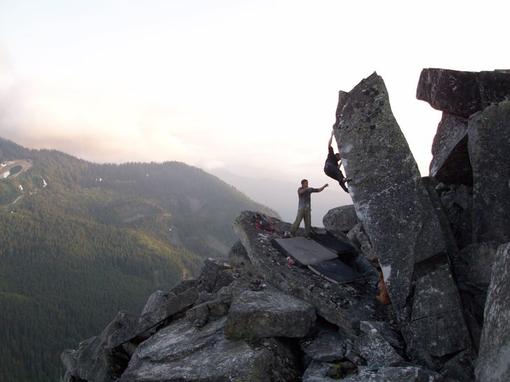 A distant shot of a climber on a boulder problem on the top of a mountain with the setting sun lighting up the fog in the background.