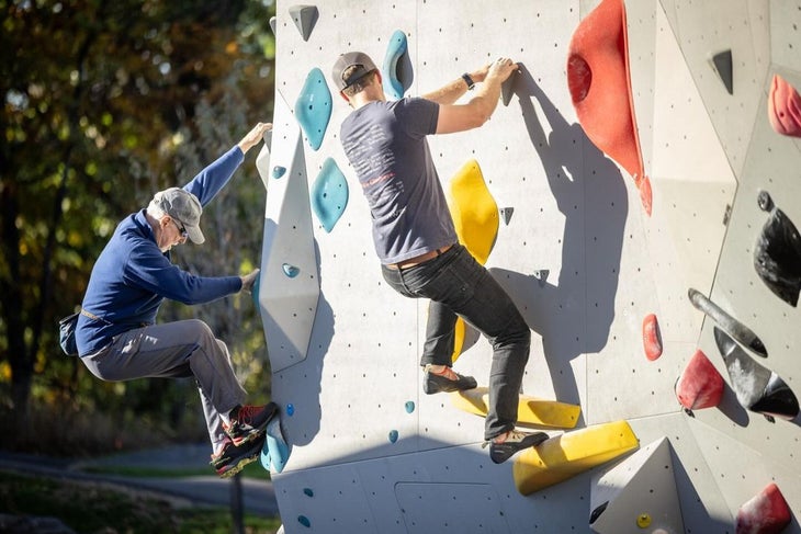 Two men climbing side by side in the Boyce Bouldering Park. One wearing climbing shoes, the other wearing approach shoes.