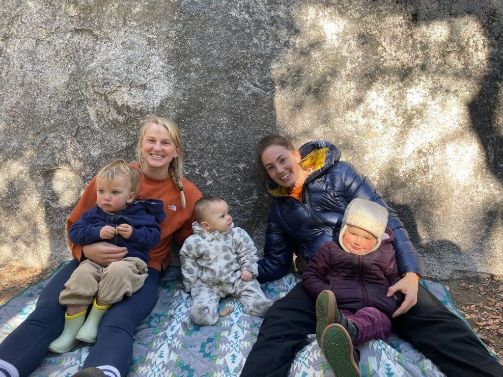two women and three little kids sitting on a blanket on the ground by a rock