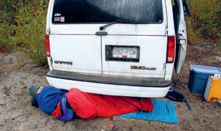 Man sleeps under a white van in red sleeping bag.