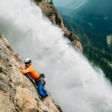 Man wearing orange climbs rock beside a waterfall wearing some of the best gift for climbers.