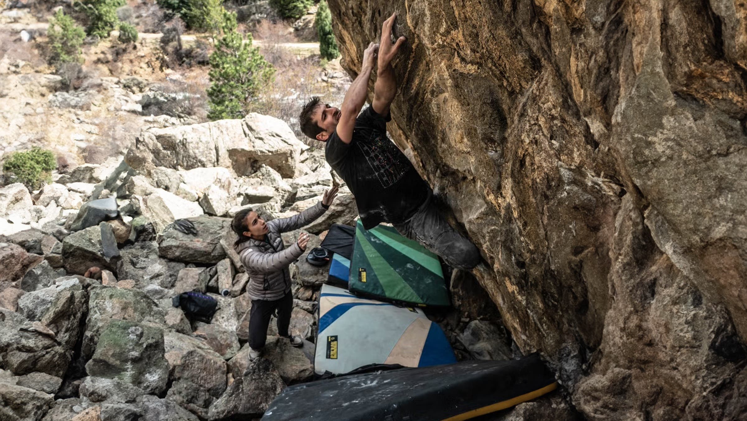 A male boulderer in a dark shirt reaches for the top of a rugged rock face while a spotter in a grey jacket guides him from below, with multiple stacked climbing crashpads positioned against the boulder in a rocky canyon setting.