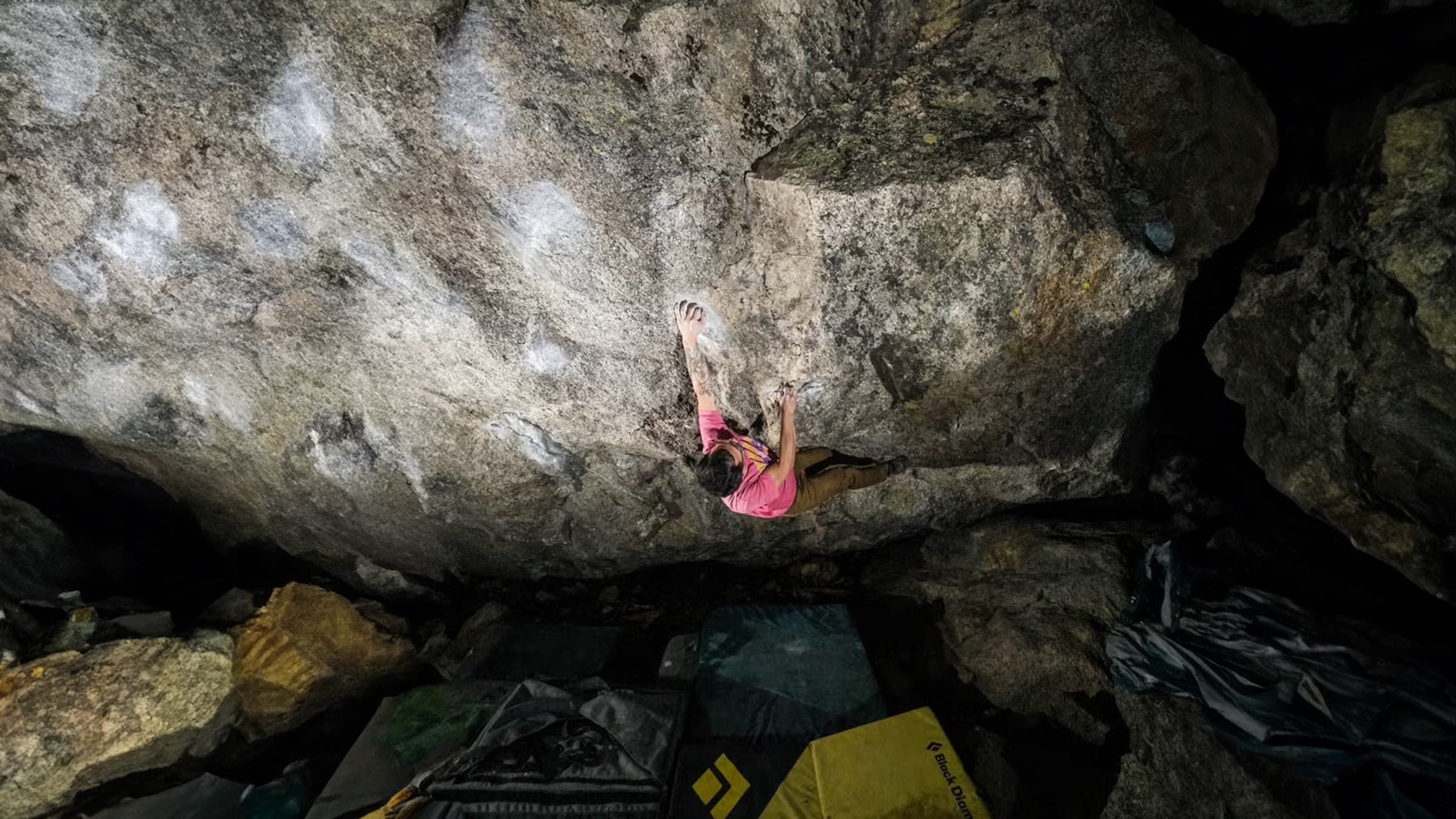 A climber in a pink shirt traverses a dramatic granite overhang at night, reaching for a chalked hold on the underside of a massive boulder, with Black Diamond climbing crashpads stacked on the rocky ground below.